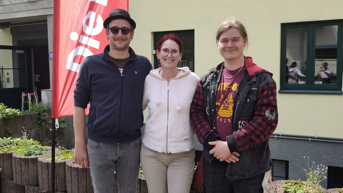 Das Foto zeigt drei Personen vor einem Haus, neben ihnen weht eine Fahne der Partei Die Linke.