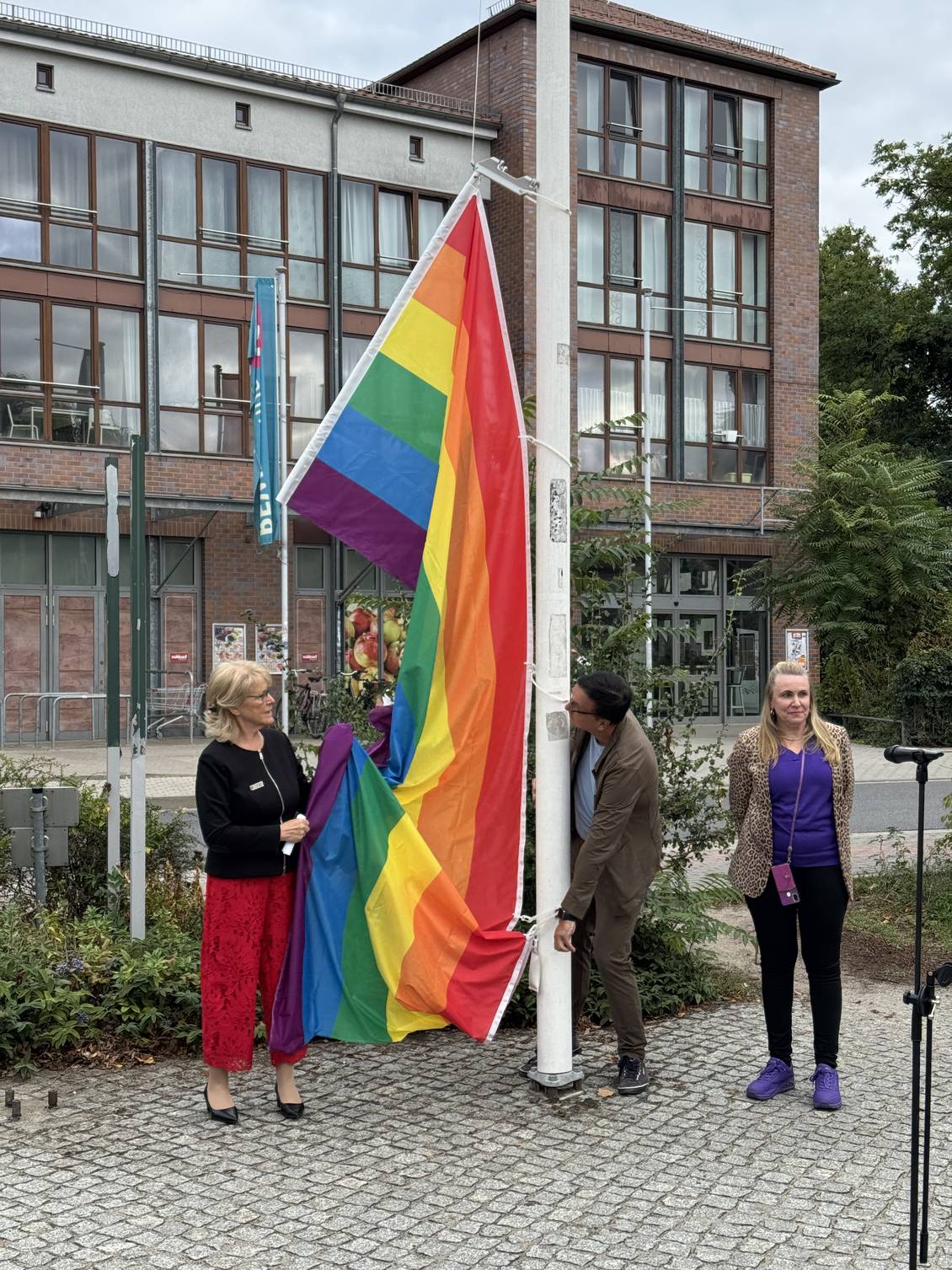 Das Foto zeigt, wie mehrere Personen an einem Fahnenmast eine Regenbogenflagge hissen.