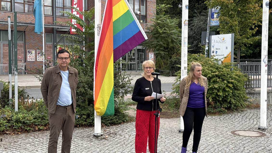 Das Foto zeigt drei Personen vor den Fahnenmasten vor dem Kreishaus in Luckenwalde. An einem Fahnenmast hängt eine Regenbogenflagge.