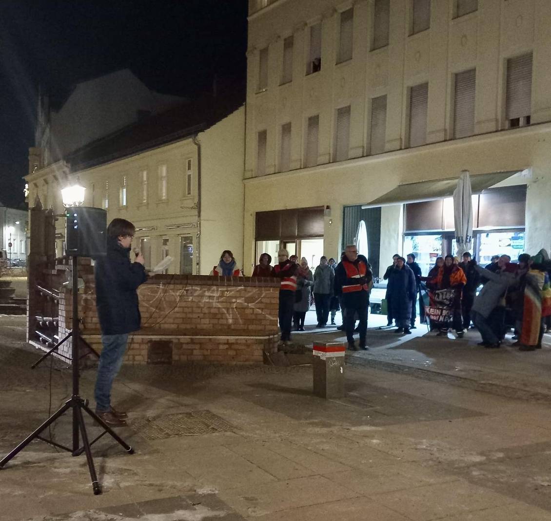 Das Foto zeigt eine Person während einer Rede, andere Menschen stehen herum und hören zu.